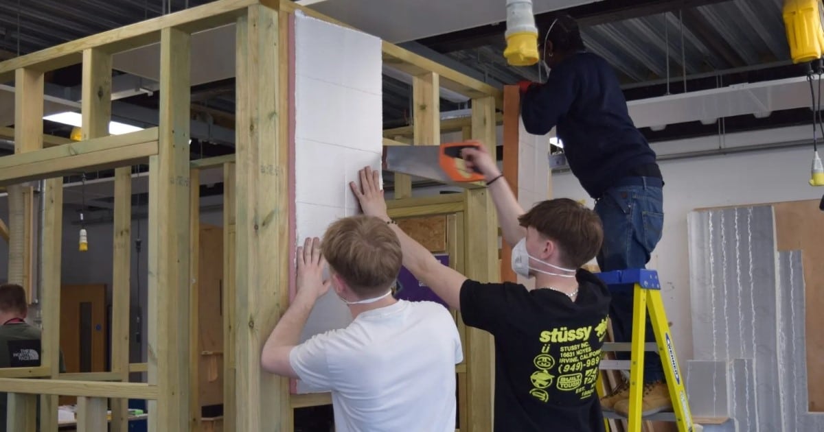 A group of three construction workers complete work on insulating a stud wall. One is stood on a yellow ladder.
