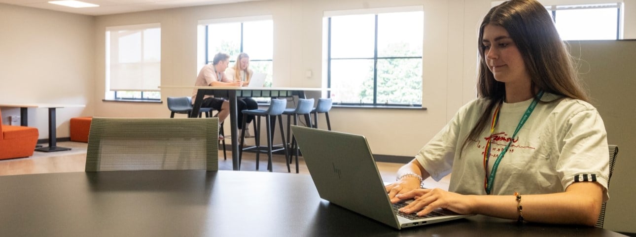 A woman in her 20s sits at a table while typing on a computer. She is in a further education campus.