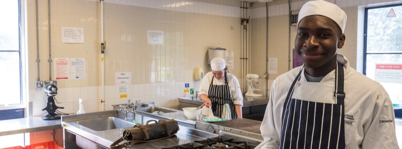A chef in a white jacket and clue apron smiles at the camera while his colleague wipes the surfaces clean.