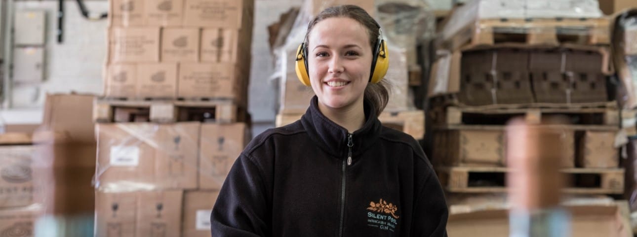 A female entry level worker in a warehouse is wearing ear protectors and a black fleece with Silent Pool Distillery written on it.