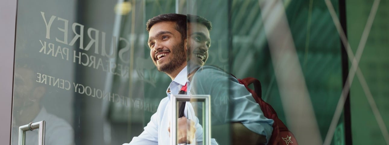 A male university student with a bag on his back walks through a glass door. It has Surrey Research Park written on it.
