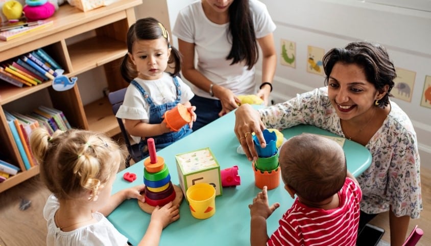 A group of young children in an early years setting sit round a table as a childcare professional talks to them.