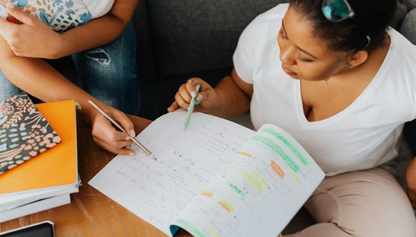 A woman in a white top is studying English with the help of a tutor. In front of her is a notebook.