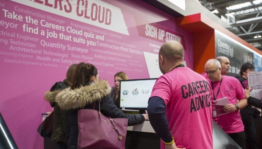 A man wearing a pink shirt with Careers Service on his back helps a woman with a computer search.