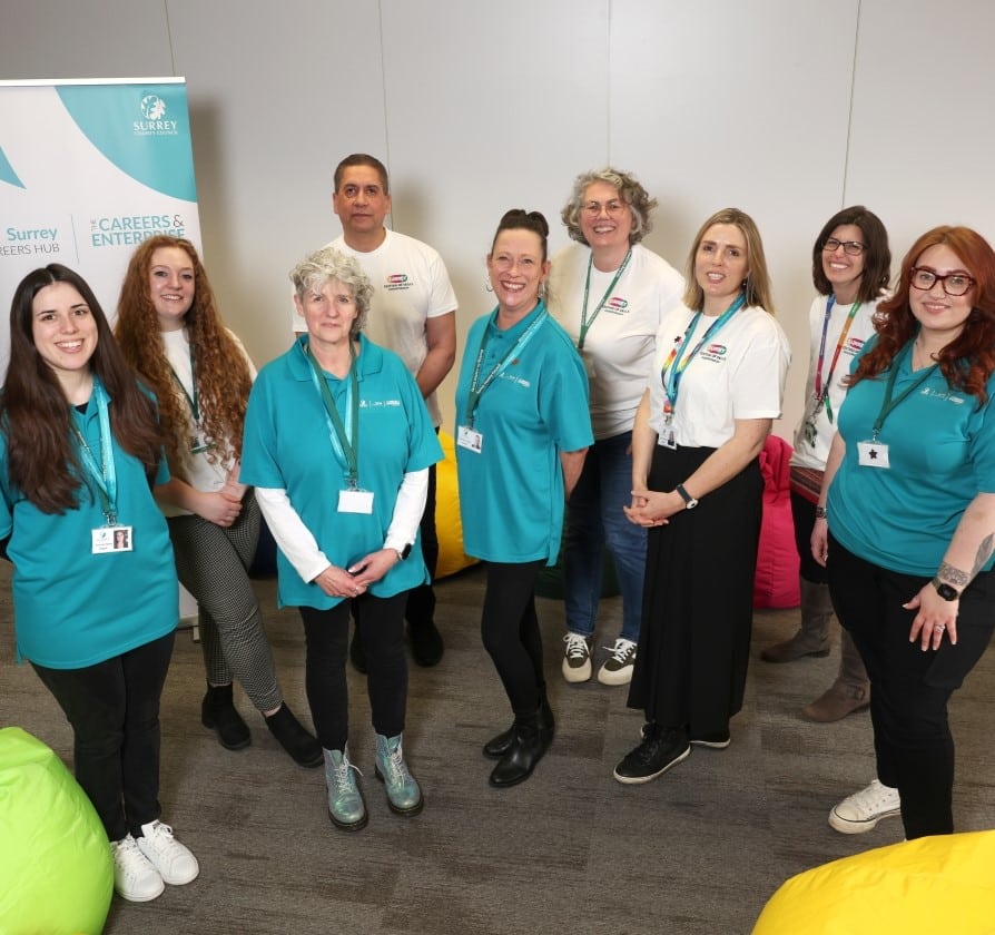 A team of professionals in a mix of white and blue tops stand in front of Surrey County Council promotional banner.