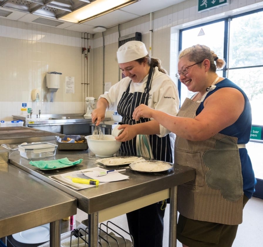 Hospitality teaching taking place at a Surrey further education campus. A tutor advises a young chef how to mix batter in an industrial kitchen.