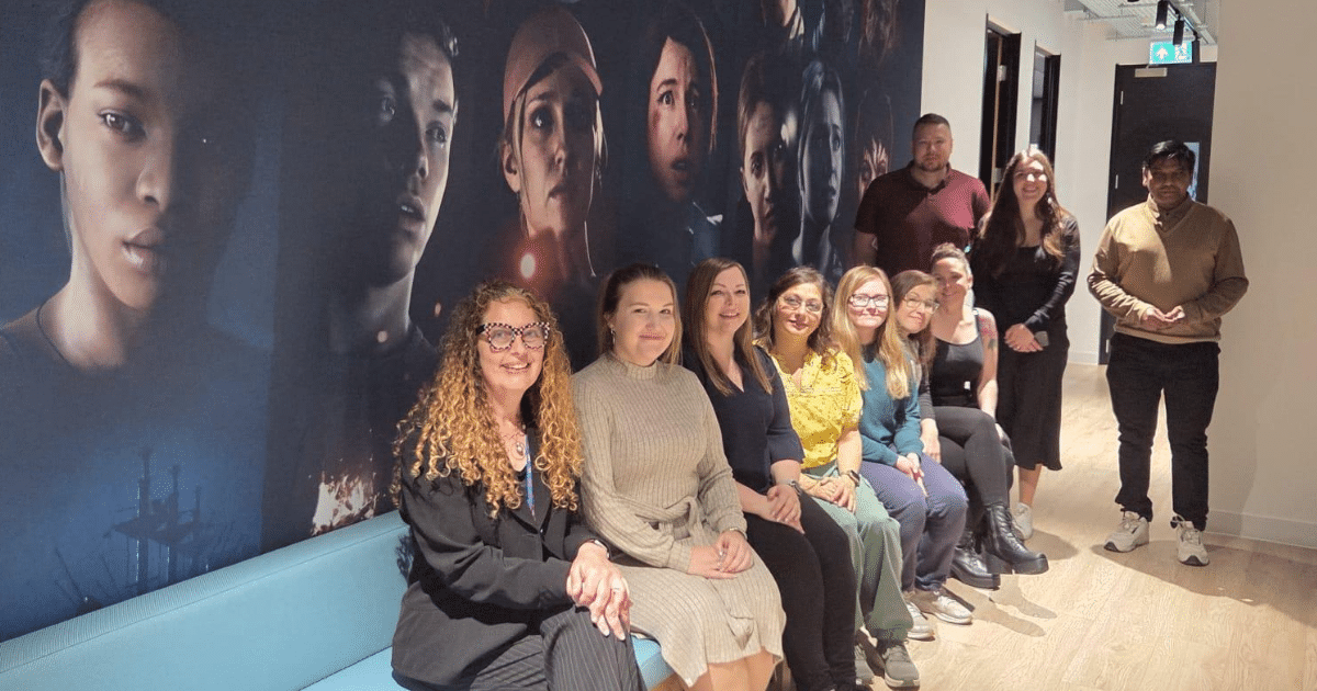 A group of people sat on a sofa smile at a camera. They are in an office with images of game developers on the wall.