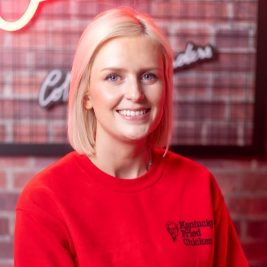 A woman with a blonde bob and red KFC UK jumper poses for a professional headshot in front of a neon red sign.