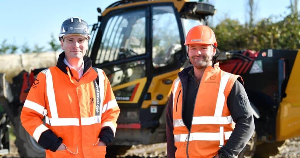 Two men in orange high vis and hard hats stand in a construction site. Behind them is a yellow plant vehicle.