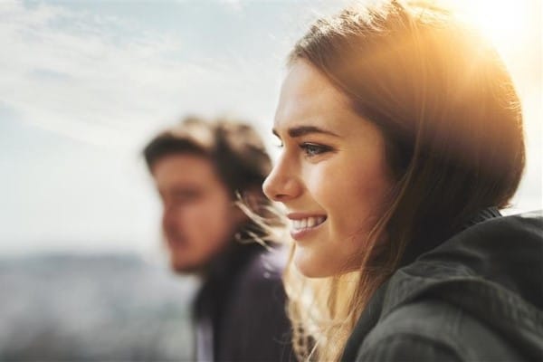 A woman with dark hair smiles as she gazes over the horizon. The sun shines through her hair.