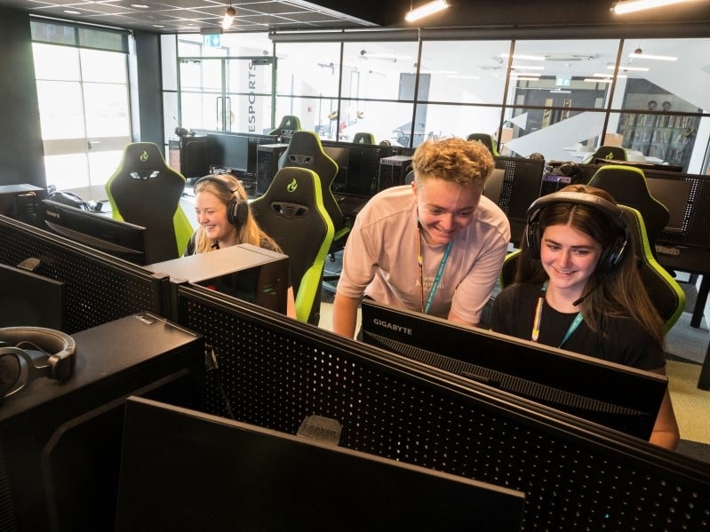 Three young people are in a games classroom learning new skills. Two are sat in black gaming chairs while one is stood looking at a screen