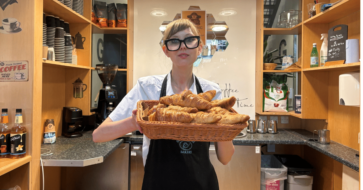 Scarlet stands in a coffee serving area and is presenting a basket of pastries | Connect to Work | STEPSurrey